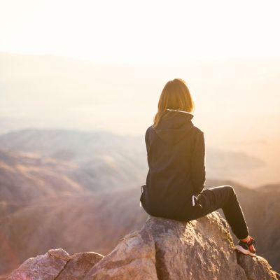 Person sitting on a large rock facing mountainous landscape at sunset.