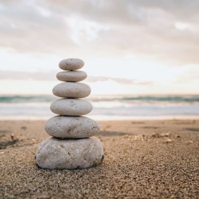 Stack of six flat stones balanced on a sandy beach with the ocean and cloudy sky in the background.